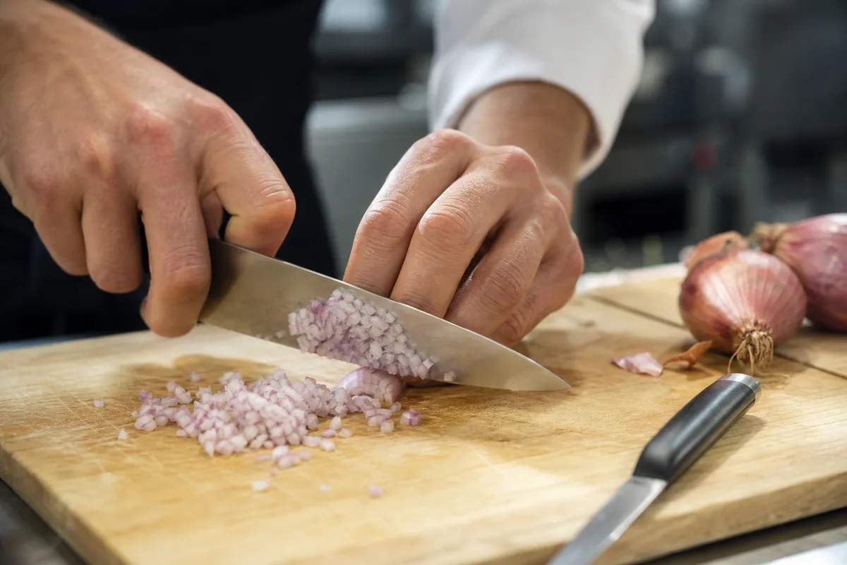 Mains de chef ciselant finement une échalote au couteau sur une planche en bois